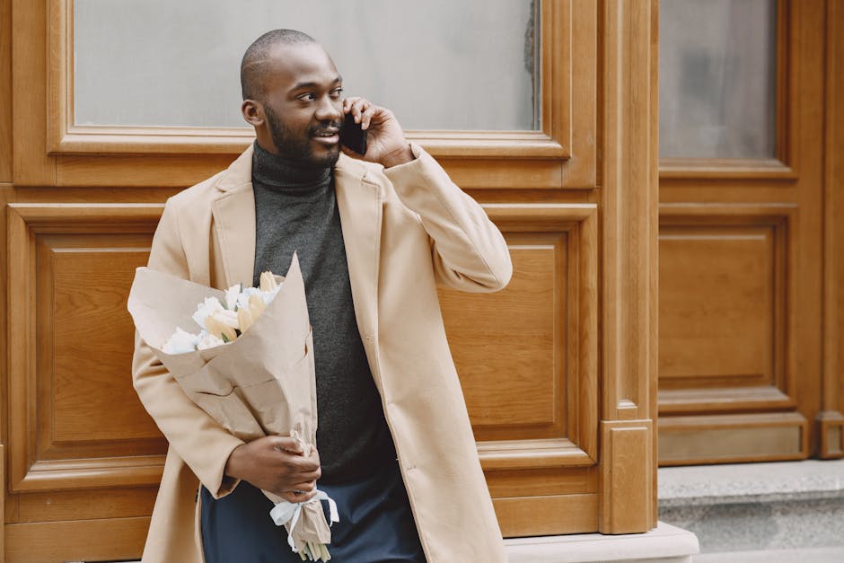A man standing outside against a wooden-paneled wall, holding a bouquet of fresh, cream-colored flowers wrapped in brown paper with white tissue paper accents. He is wearing a beige overcoat layered o