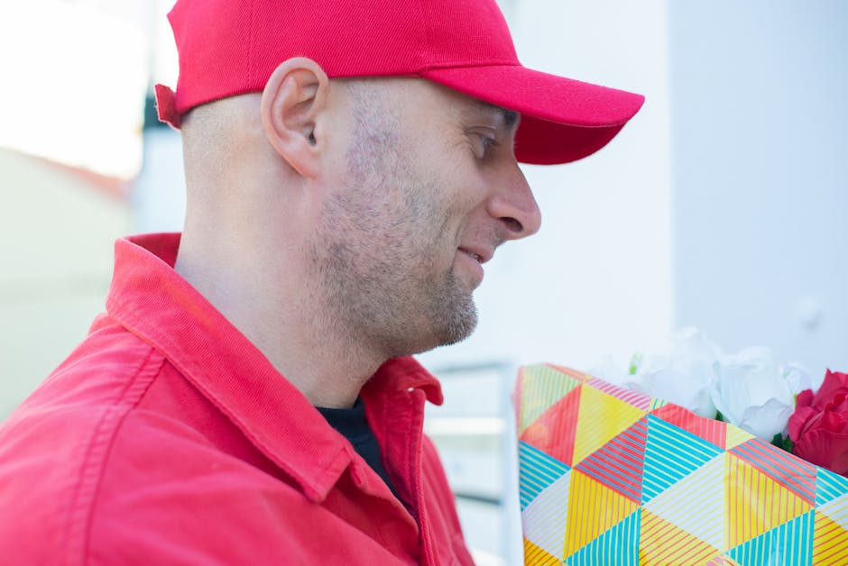 A man in a red baseball cap and matching red jacket holds a brightly wrapped floral gift, likely a bouquet of fresh flowers, with a geometric patterned wrapping paper featuring colors such as blue, ye