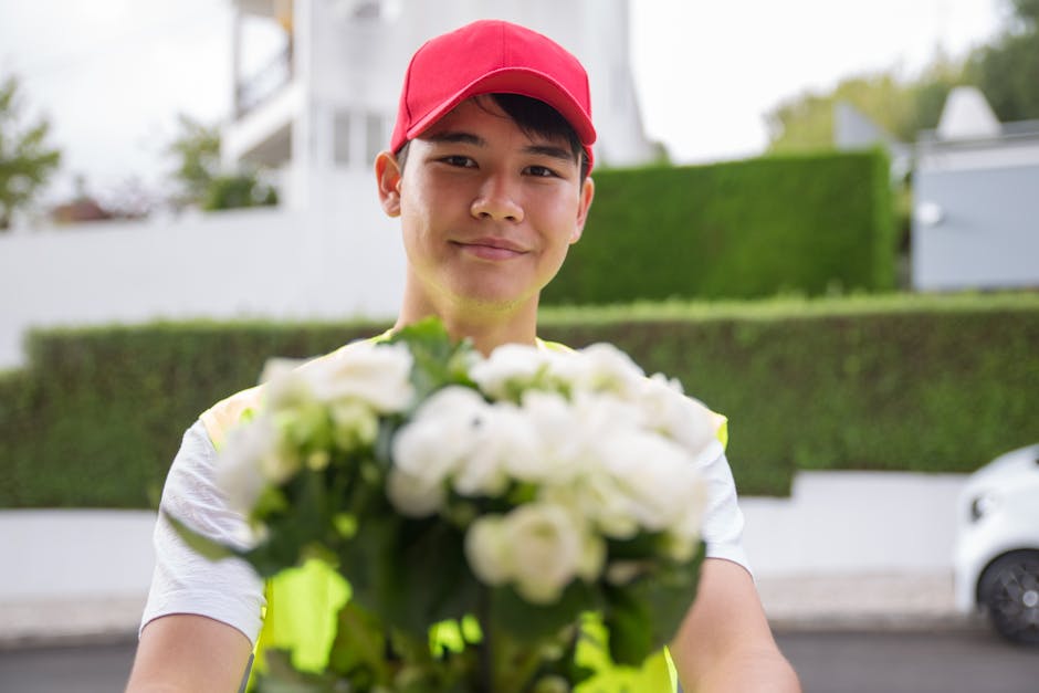 A young male flower delivery courier wearing a red cap and a neon yellow vest is standing outdoors in a suburban area, holding a bouquet of white flowers with a mix of green foliage. The bouquet appea