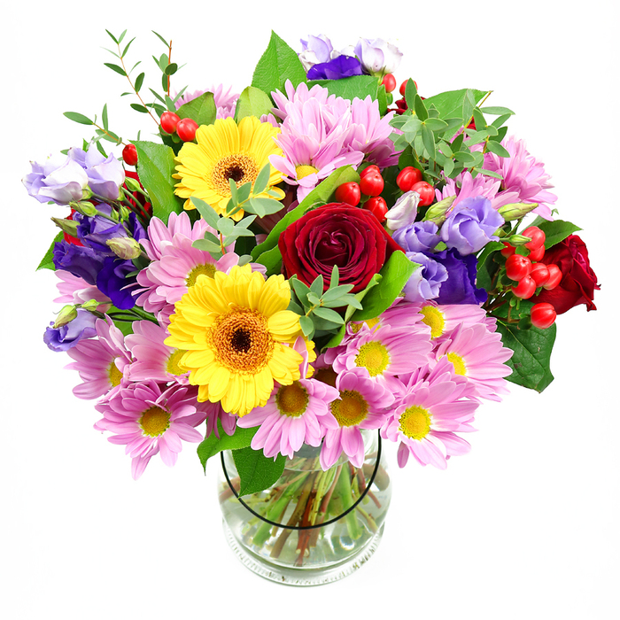 An overflowing mixed bouquet stands proudly in a cylindrical glass vase, its colours vivid against a crisp white backdrop, as though ready to be delivered to a doorstep in Redbridge. At the heart of the design, rich red roses unfurl velvety petals in tight spirals, giving depth and a sense of timeless romance. Framing these are two radiant yellow gerbera daisies, each bloom with a wide circle of golden petals encasing a dark, brown-orange centre that adds definition and contrast. Surrounding this central cluster, generous sprays of soft pink daisies or chrysanthemums create a cushion of pastel colour; their many fine petals and tiny yellow centres bring a sense of gentle charm and fullness, like a flowering border in a well-kept local garden. Threaded thoughtfully throughout are lisianthus stems in tones of purple and lavender, their graceful, bell-shaped blossoms and slender buds adding height, coolness, and a touch of sophistication. Among the petals, bright red Hypericum berries appear in small, rounded clusters, their smooth, shiny surfaces catching the light and introducing interesting texture. The entire arrangement is bound together by layers of greenery: broad, dark green leaves provide structure, while more delicate, light green sprigs and shoots lend freshness and movement, echoing the foliage you might notice walking through Wanstead Park. The stems are clearly visible through the transparent vase, neatly cut at equal lengths and submerged in clear water that suggests just-picked freshness. The bouquet feels joyful yet considered, equally at home as a birthday surprise, a thank-you gift for a neighbour, or a colourful centrepiece for a family gathering in Redbridge, radiating warmth and celebration.